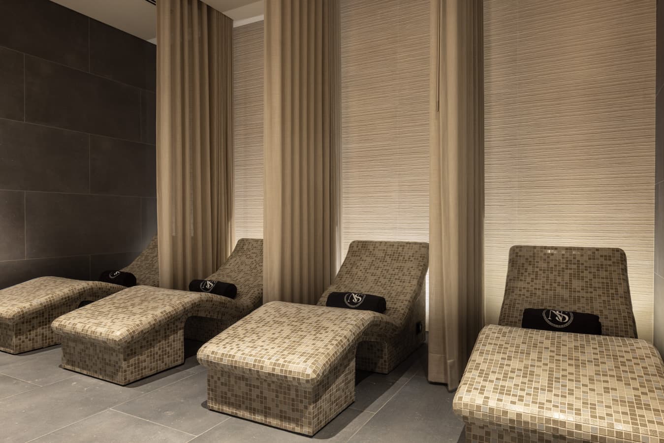 Row of heated mosaic relaxation loungers with rolled towels, separated by linen curtains