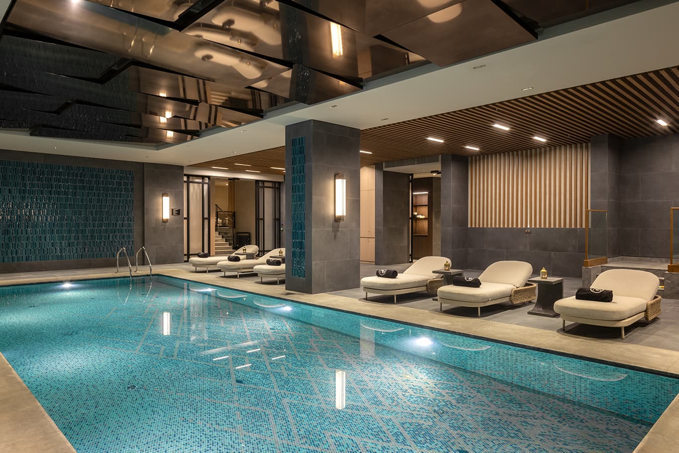 Wide view of the indoor pool hall with loungers, wood-slat ceiling and brass sconces