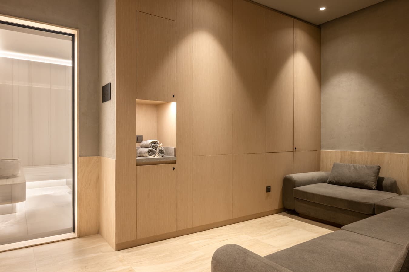 Wood-panelled lounge with a grey sofa and a lit towel niche beside the glass door to the thermal area